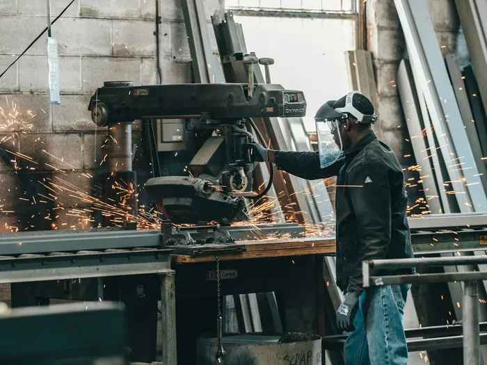 Pracownik w przyłbicy tnie metal na hali produkcyjnej, wokół iskry